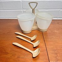 Front view of vintage plastic Tupperware condiment caddy with three containers and three ladle spoons on wood table against white brick wall
