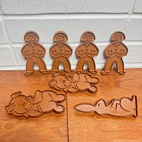 Photo showing four human-shaped cookie cutters standing upright and three animal-shaped cutters (two pigs and one rabbit) laid flat on a wooden surface with a white brick wall background.