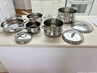 Set of 8 stainless steel pots and pans with matching lids displayed on kitchen counter, showing different sizes and types including frying pans and saucepans.