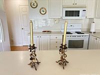 Pair of gold-toned metal candle holders on kitchen counter with taper candles inserted showing front view and overall size.