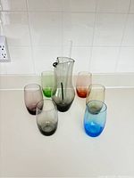 Photo showing six colored glasses arranged around a decanter with an internal stirrer on a white counter and white tiled wall background.