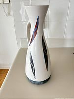 Front view of the white pottery vase with leaf decoration and curved handle on a light-colored surface with white tiled backsplash.