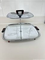 Front angle view of the chrome two-tier tray showing the dark red Bakelite handle on the top tier and the larger bottom tier with side handles.