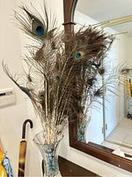 Bundle of natural peacock feathers displayed in a decorative clear glass vase in front of a mirror. Feathers show the distinctive blue-green 'eye' pattern of peacock feathers.