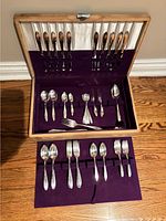 Wooden box opened displaying silverware including knives, spoons, forks, and serving utensils arranged in slots.
