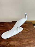 Side view of the white porcelain peacock figurine on a wooden surface, showing sleek and elongated design with detailed tail.