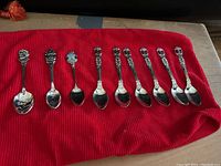 Full view of nine souvenir spoons laid out on a red cloth. Each spoon has a distinct decorative top representing different places or themes.
