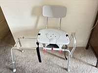 Photo of swivel sliding transfer bench showing white plastic seat, grey padded backrest, metal frame with safety straps, and rubber-tipped legs.