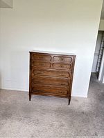 Front view of a medium brown solid wood five drawer dresser with metal handles, carved detailing and tapered legs, standing on carpet against a white wall.