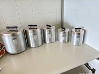 Five vintage aluminum canisters labeled for flour, sugar, coffee, grease, and tea, arranged in size order on a kitchen counter.