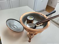 Copper and brass three-legged chafing dish with lid removed, showing interior including the wick holder and wooden-handled spoon inside the dish.