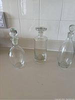 Set of three clear glass decanters with stoppers arranged side by side against a white tiled background, showing their various shapes and sizes.