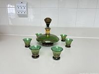 Full lot of one decanter with stopper and six matching shot glasses, showing green glass with gold stripes on white countertop