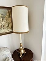 Single lamp showing full height with tall cream shade and ornate gold-accented metal base on dark wood platform.