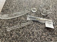 Photo shows multiple clear crystalware items on granite countertop including a rectangular glass dish with three compartments, two glasses (one tumbler, one mug), two cutter rests, and a new packaged cutting utensil set.