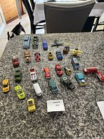 Overview of 26 preowned diecast toy vehicles and figurines in assorted colors and styles on granite table surface.