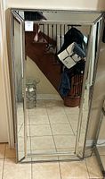 Full view of the large rectangular floor standing mirror leaning against the wall showing the beveled glass edges and metal frame with hanging attachments on back.