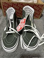 Pair of Vans SK8 Hi green high-top sneakers showing front and top views with white laces and tags, placed on grey surface with Vans box behind.