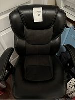 Front view showing a black office chair with cushioned backrest and leatherette and mesh upholstery. White stitch detailing visible. Chair is positioned near white door and some bags on side.