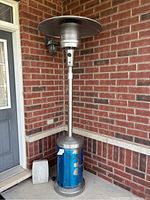 Full view of the outdoor propane patio heater showing tall metal pole with wide top heat reflector, blue protective film on propane tank casing, and weathered surface.