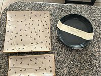 Two square ceramic plates with gold edges and black abstract markings, lying flat on a granite countertop beside a stack of four black round plates wrapped with a Katie Kime label.