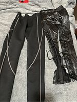 Photo showing two pairs of black pants: sauna sweat pants with reflective seam detailing on the left, faux leather lace-up leggings on the right, both laid flat on a grey blanket.