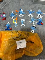 Eleven Smurfs plastic figures laid out on gray fabric showing variety and condition.