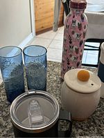 First photo showing blue glass drinking glasses, pink dog-print water bottle, ceramic container with yellow knob, and black travel mug