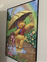 Side angle showing the framed Winnie the Pooh poster featuring Pooh with an umbrella and Piglet walking beside him in a puddle, with a bright rainbow and flowers in the background.