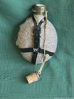 Front view of the Czech Army water bottle showing the gray wool cover with black leather straps and cork plug stopper attached by a rope.