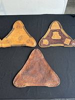 Top view of three hand-tooled leather tripod stool seat saddles arranged on black background, showing tooling and edge stitching.
