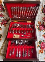 Photo of full set of flatware laid inside the open wooden chest showing three tiers containing forks, spoons, knives, and serving pieces with intricate floral designs on handles