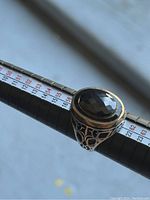 Close-up front view of the men's silver ring on a measuring tool showing the large faceted black oval stone and filigree silver band.
