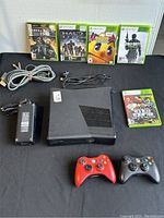 Xbox 360 console, power adapter, AV cable, two controllers (red and black), and five Xbox 360 games arranged on black surface.