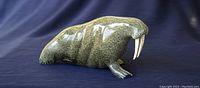 Front-right three-quarter view of green-speckled soapstone walrus carving with white tusks on blue backdrop