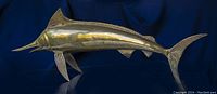 Side view of large brass swordfish sculpture showing detailed fins and elongated body shape on blue background.