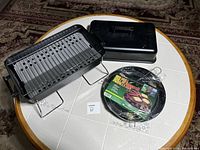 Both portable charcoal BBQ grills displayed, one open showing charcoal tray and another closed with lid. Accessories including pan and utensils packaged together.