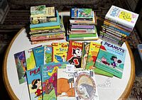 Wide shot of multiple stacks of children's books piled on a table, showing a variety of titles including Peanuts coloring books and classic fairy tales.
