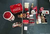 Full display of Coca-Cola collectible lot with cookie jars, tins, napkin dispenser, coasters, trinket tray, and other branded items shown on black background.