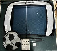 Photo showing the pop-up soccer goal frame in black and white mesh with Franklin branding, a soccer ball shaped carrying case, instruction sheet, and metal support rods laid out on floor.