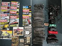Full overview of lot with curved track pieces stacked vertically, several transformers/power packs with wiring, multiple O Scale train cars in green, red, and black, and magazines/brochures laid out on left side.