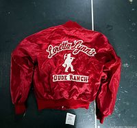 Full view of the back of the red satin jacket showing large white embroidered text 'Loretta Lynn's Dude Ranch' with cowboy silhouette graphic.