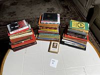 Three stacks of assorted books displayed on a white table. Titles and authors include Shirley MacLaine, Pearl S. Buck, and others. Books vary in size and color.