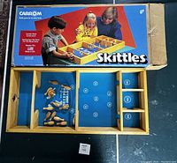 Photo showing Carrom brand original Skittles game box and wooden game board with pins and spinning top inside.