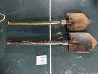 Two WWII folding entrenching shovels laid side by side on a dark surface, showing length and folding mechanism