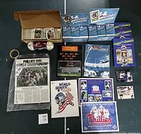 Full overview of the sports collectibles including sealed football card boxes, loose baseball cards, sealed baseball, Phillies World Championship plaque, magazines and programs arranged on floor.