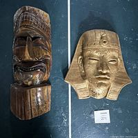 Two vintage wall hangings, tiki mask and King Tut mask, shown together on dark floor.