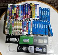 Wide view of the VHS tape lot showing approximately 5 rows of tapes on a table, titles visible on spines including animated Disney and Warner Bros movies and cartoon compilations. Multiple labels on tapes are visible.