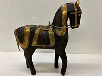 Front and side view of dark wooden horse standing on four legs, adorned with gold-tone metal accent bands on saddle, mane, tail, and body.