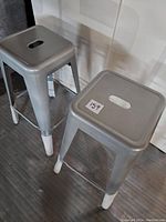 Two silver metal bar stools with white plastic or rubber caps on the legs, showing a side angle revealing the cut-out handle on the seat top
